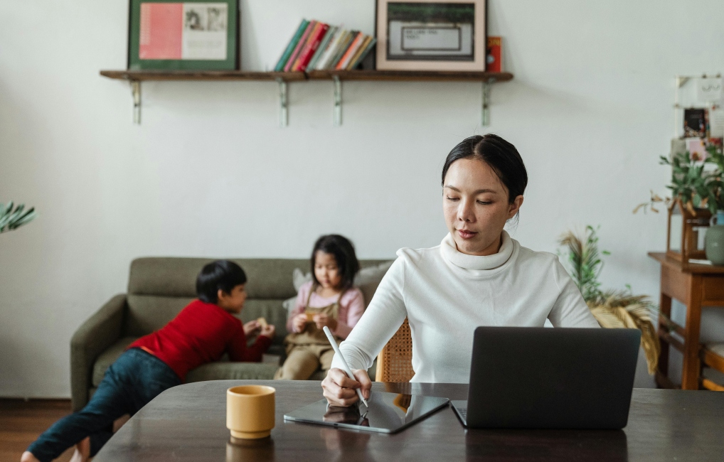 Mother working at a desk while two children are playing behind her on a sofa
